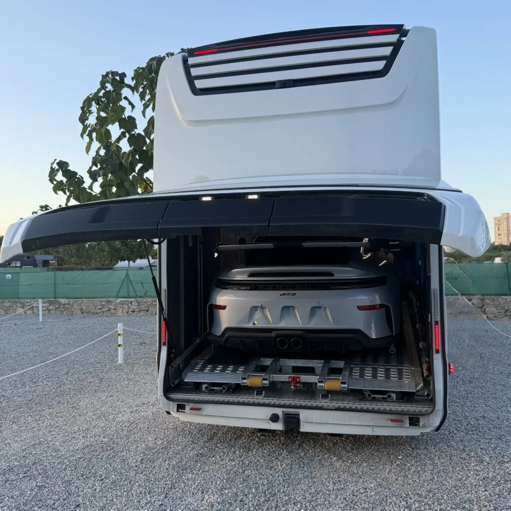 Liner-Wohnmobil mit geöffneter Heckgarage und einem Porsche darin, auf einem XXL-Stellplatz im Sunset Camper Park